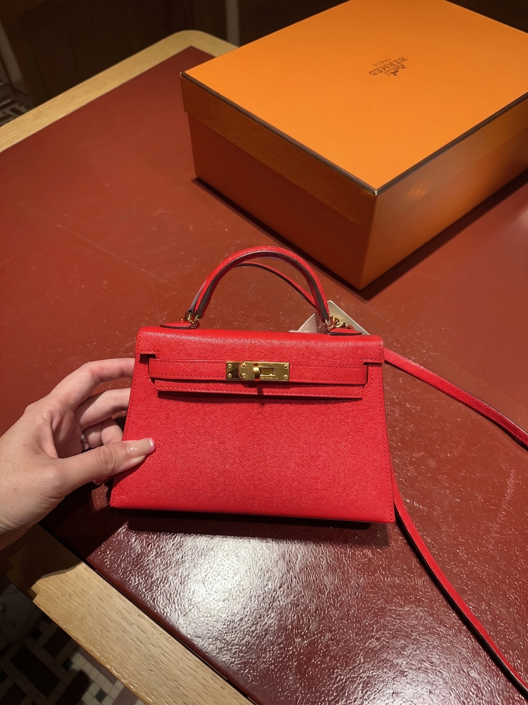 Hand holding a red Hermès Mini Kelly Sellier with gold-tone hardware and shoulder strap