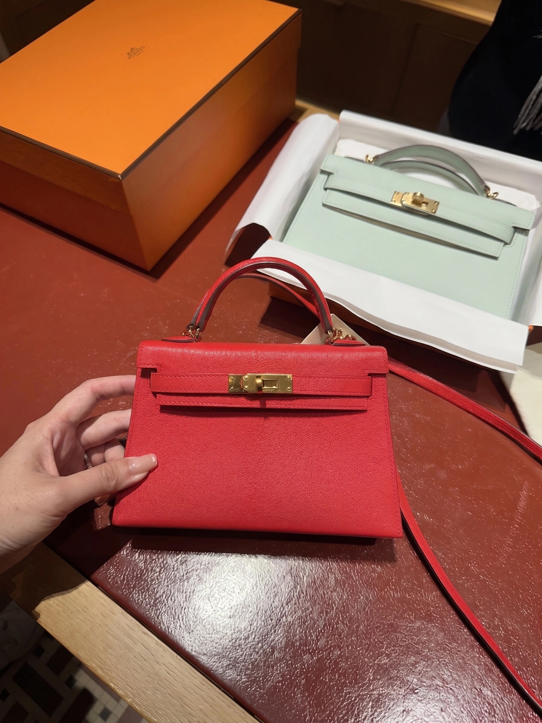 Red Hermès Mini Kelly held in front of a pale green Kelly To Go wallet for size comparison